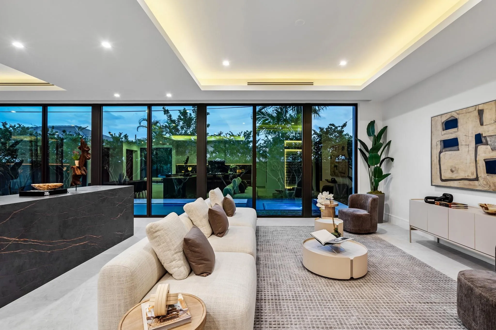 A stunning contemporary living room highlighted by sleek architectural lines, recessed ceiling lighting, and expansive walls of glass overlooking lush greenery. Sophisticated and serene, this open space seamlessly blends indoor elegance with effortless outdoor connection.