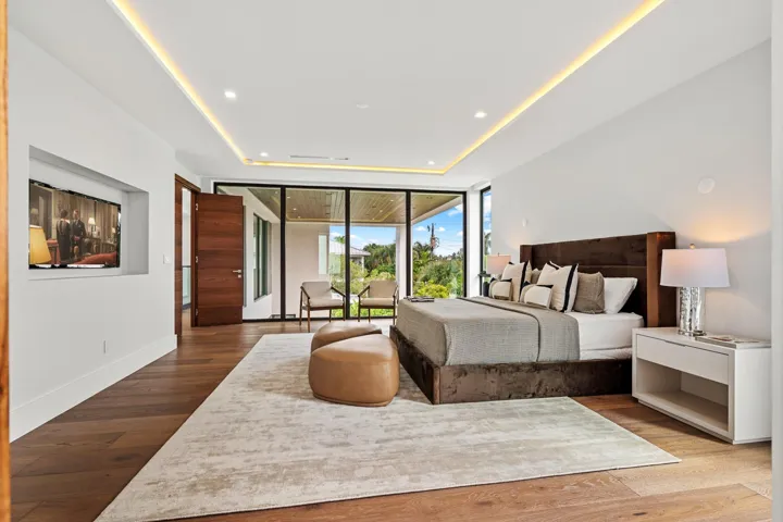 A luxurious modern bedroom retreat featuring warm wood flooring, a statement upholstered bed, and seamless access to a private terrace beyond. Clean architectural lines and expansive glass create a serene, light-filled sanctuary designed for elevated comfort.