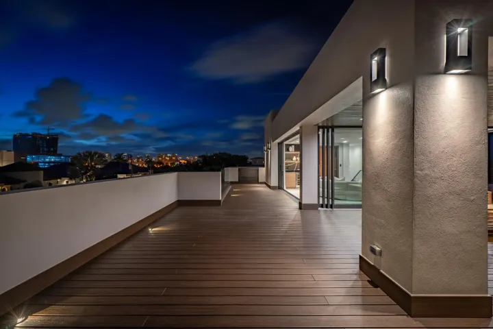 A sleek upper-level terrace illuminated by modern wall sconces, set against the dramatic glow of the evening sky. Expansive wood decking and clean architectural lines create a sophisticated outdoor retreat designed for elevated nighttime entertaining.