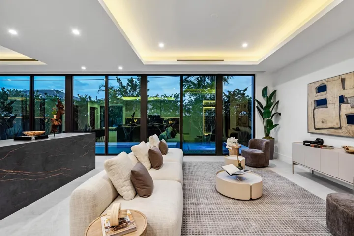 A stunning contemporary living room highlighted by sleek architectural lines, recessed ceiling lighting, and expansive walls of glass overlooking lush greenery. Sophisticated and serene, this open space seamlessly blends indoor elegance with effortless outdoor connection.
