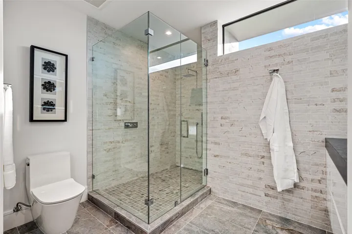 A bright, modern bath showcasing a glass-enclosed walk-in shower, sleek fixtures, and crisp neutral stone finishes. Clean-lined and thoughtfully appointed, this spa-inspired space offers comfort with contemporary sophistication.