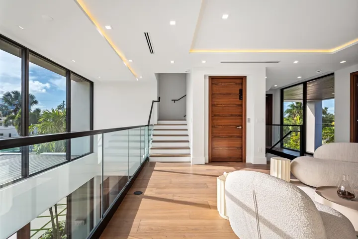 A striking upper-level gallery hallway featuring warm wood floors, sleek glass railings, and expansive windows that flood the space with natural light. Clean architectural lines and contemporary finishes create a sophisticated transition between the home’s private retreats.