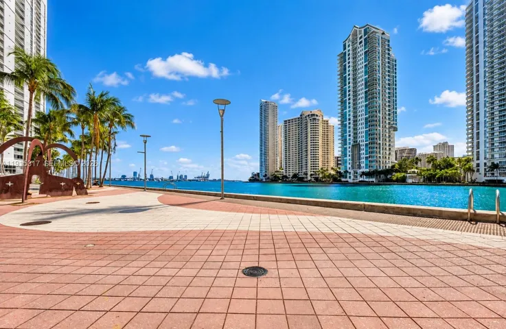 Waterfront promenade featuring a patterned paver walkway, mature palm trees, contemporary streetlights, and a metal art installation