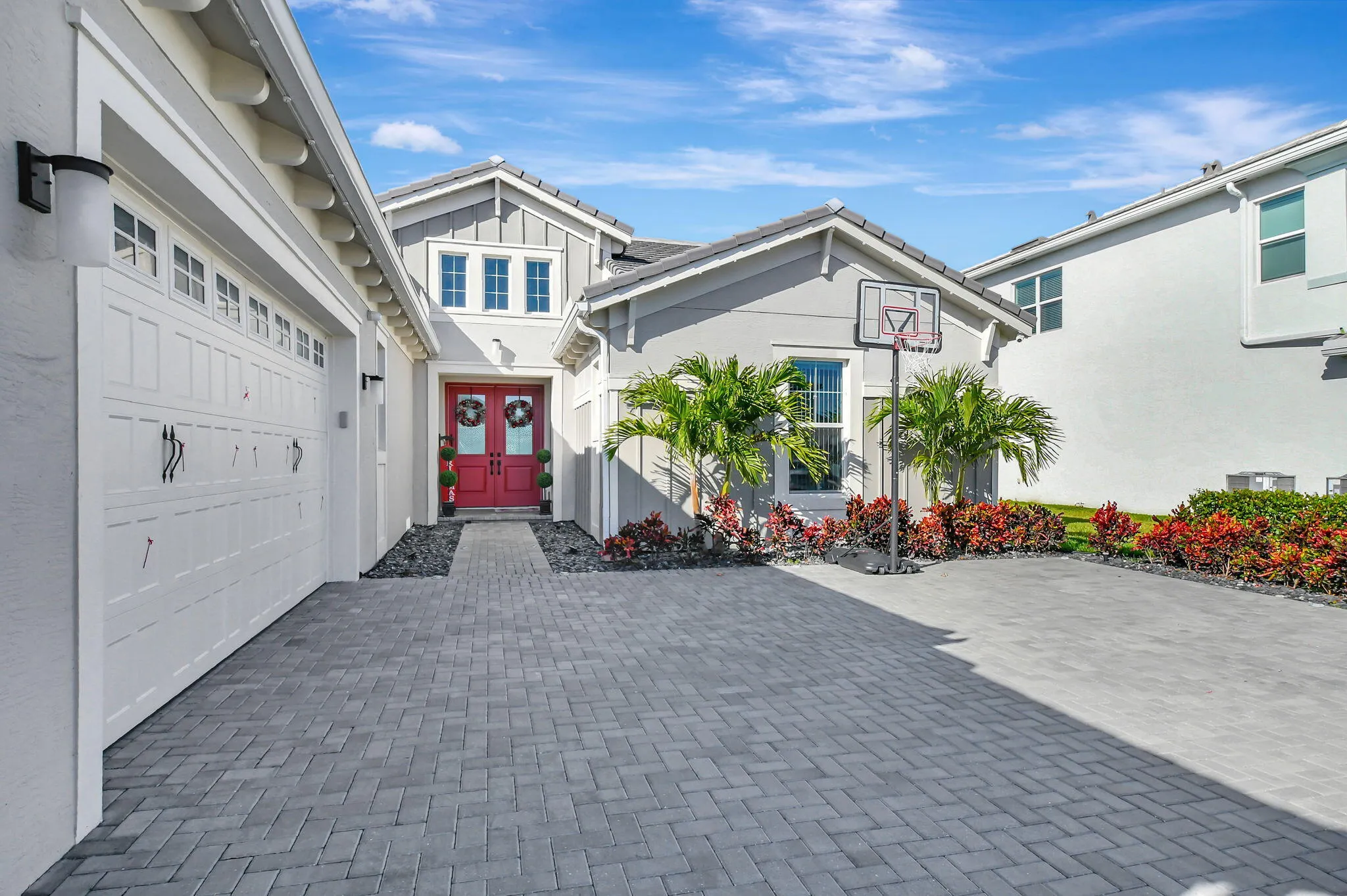 Exterior featuring a gray stucco finish with white trim, a red double-door entry, a two-car garage with paneling, a paver driveway, and varied landscaping