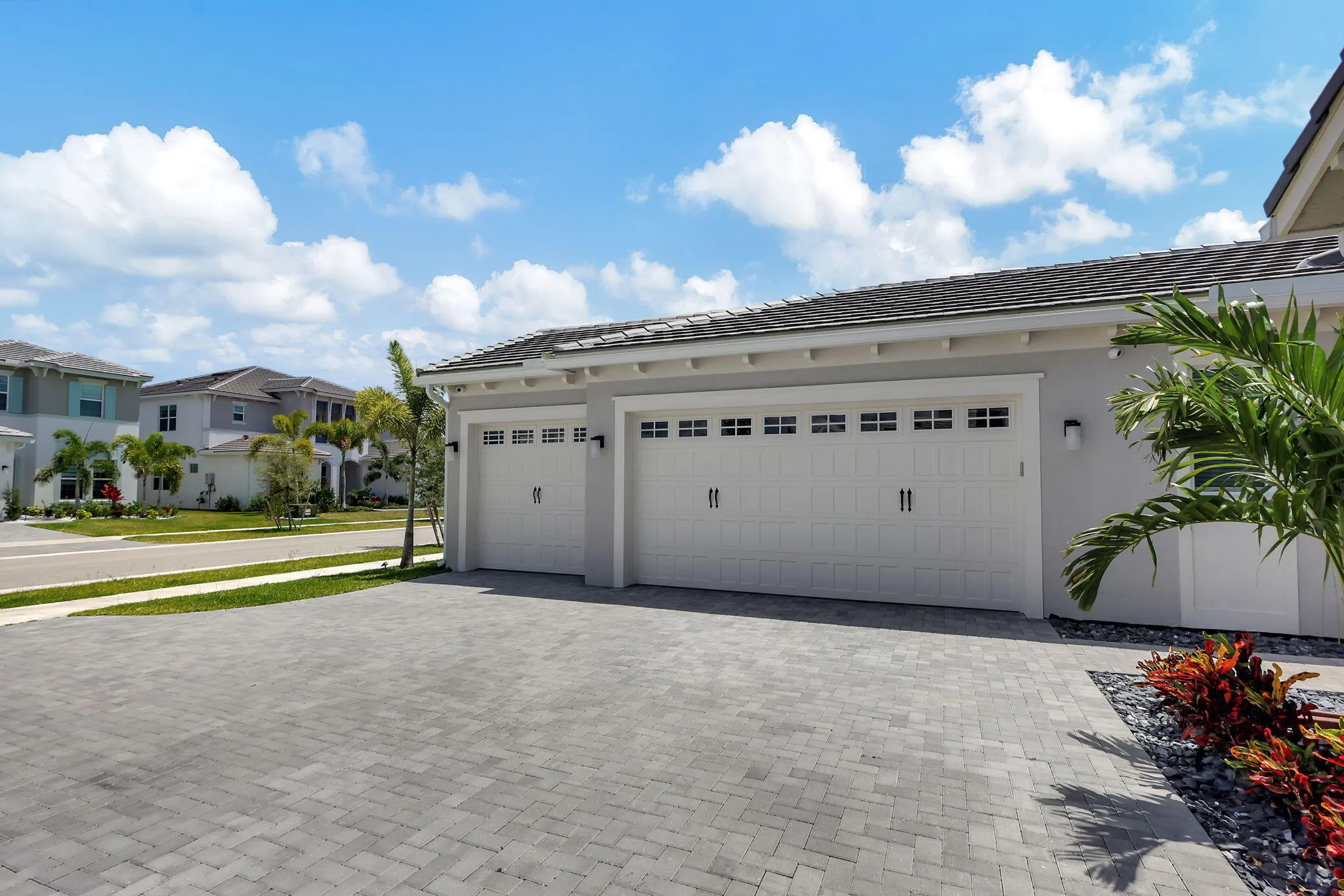 Paver driveway, dual garage doors with window accents, exterior wall sconces, tile roof, and landscape plantings