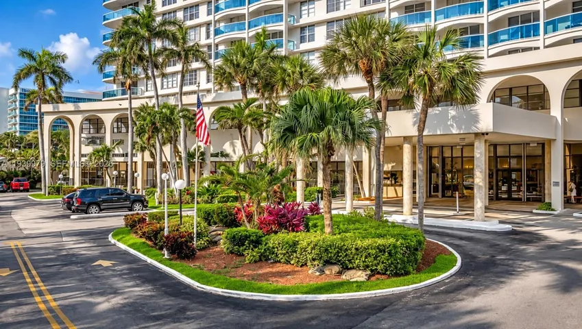 Multi-story residential building exterior featuring a porte-cochere entrance, arched structural supports, glass balcony railings, and a landscaped circular driveway with mature palm trees