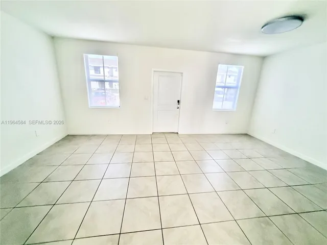 Open-concept room featuring light-toned tile flooring, neutral walls, two large windows, a central paneled entry door, and flush-mount ceiling lighting