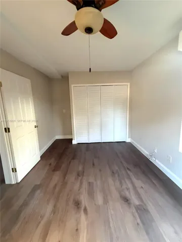 Room featuring wood-finish flooring, white louvered-panel closet doors, a multi-panel interior door, and a ceiling fan with integrated lighting