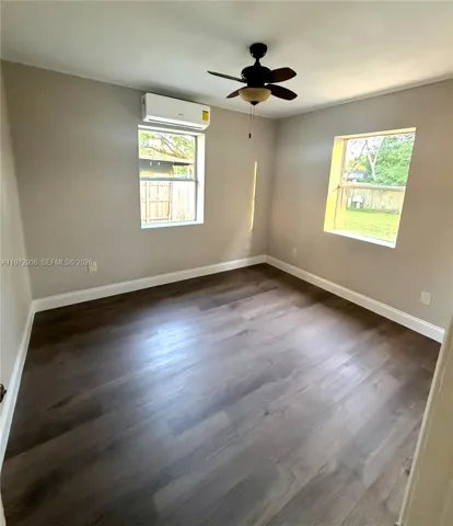 Room featuring wood-finish flooring, two windows, wall-mounted air conditioning unit, and ceiling fan with integrated light fixture