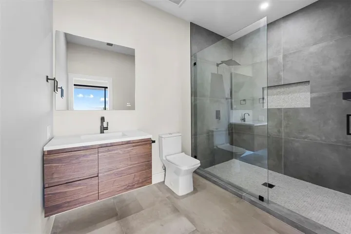 Floating wood-finish vanity with an integrated basin, frameless mirror, walk-in shower with glass enclosure, large format tile shower walls, and a mosaic tile shower floor