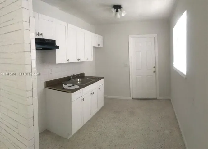 Kitchen area featuring white shaker-style cabinetry, dark laminate countertops, stainless steel sink, and exposed painted brick accent wall