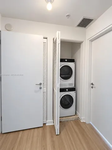 Stacked laundry unit housed within a closet featuring white louvered doors, set against wood-finish flooring