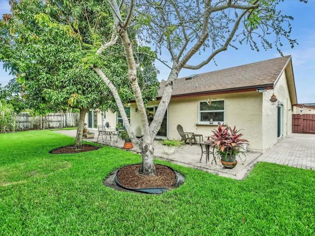Fenced backyard and side yard view with green lawn, patio area, mature tree coverage, and exterior access around the home.