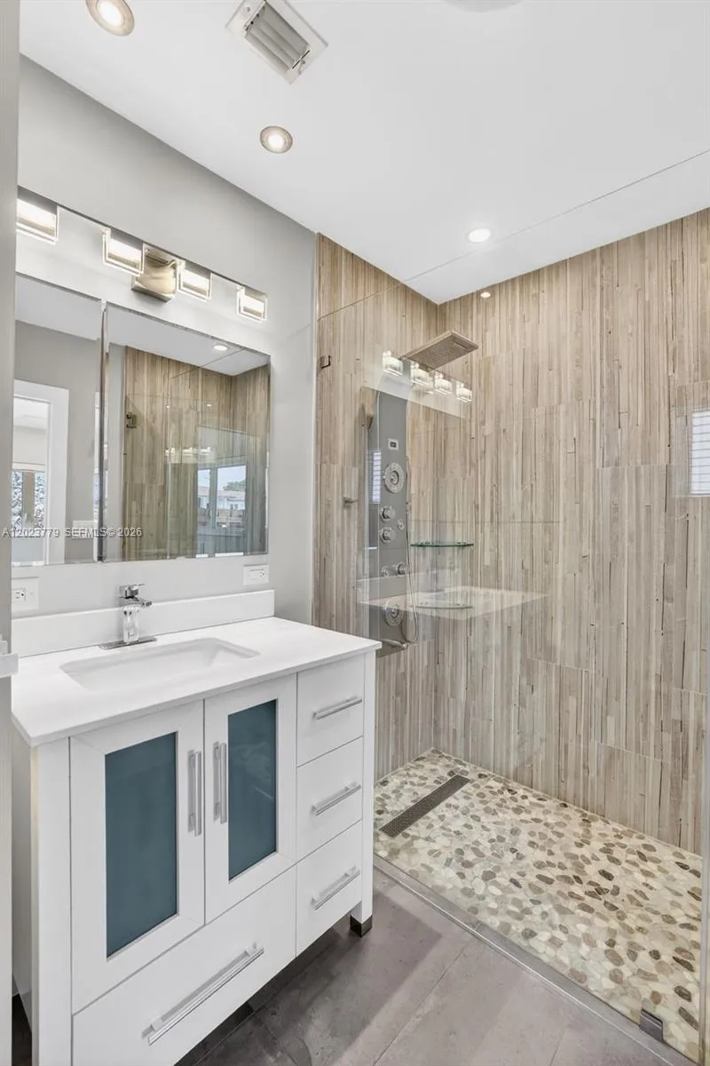 Primary bathroom w/ frameless glass enclosure, river rock look tile, floor to ceiling wood look plank tile, medicine cabinet and upgraded vanity and flooring