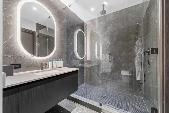 Modern floating vanity with a light-toned countertop, integrated oval LED-backlit mirror, walk-in glass shower enclosure, large-format grey stone-patterned wall tiling, and pebble-texture shower flooring