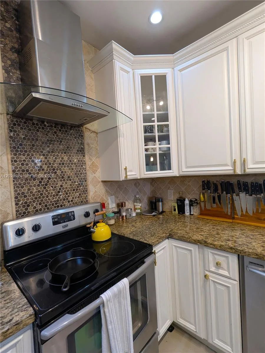 Updated cooking area featuring a stainless-steel range, modern hood, granite countertops, and mosaic tile backsplash, complemented by well-kept cabinetry and organized storage.