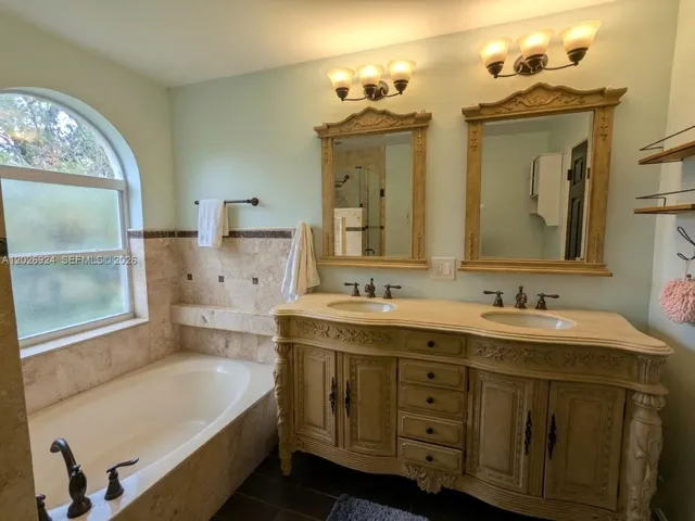 Elegant primary bathroom featuring a large soaking tub with marble tile surround, frosted arched window, glass door shower and a double-sink vanity with matching framed mirrors and classic bronze fixtures.