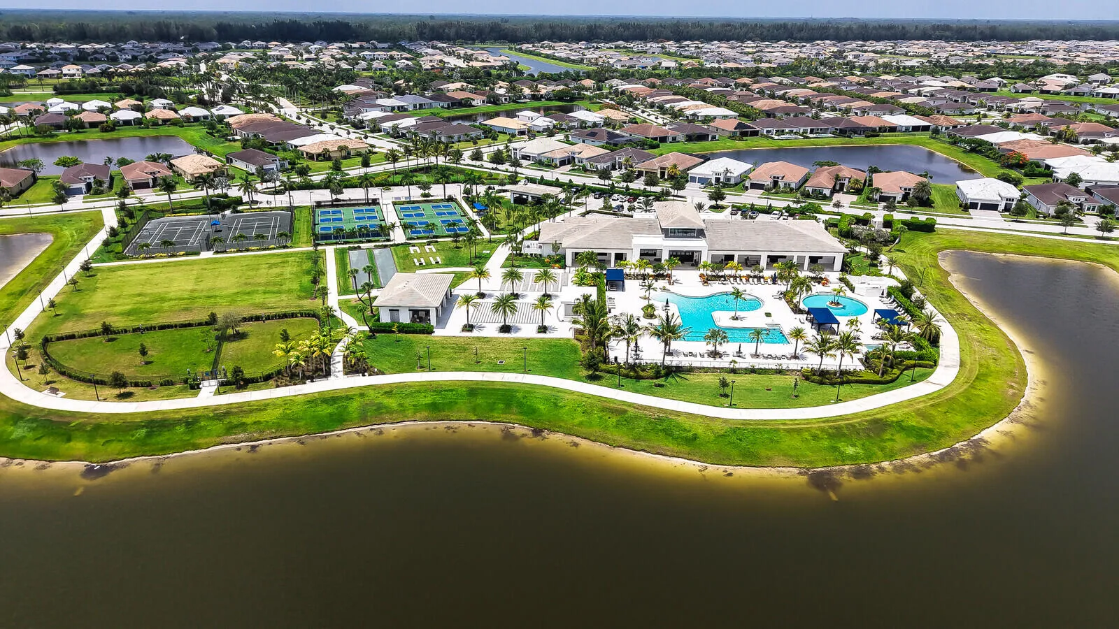 Aerial view showcasing community amenities featuring a large central clubhouse, expansive swimming pool with integrated sun shelves, multiple tennis courts, pickleball courts, and extensive manicured green spaces bordering a substantial body of water