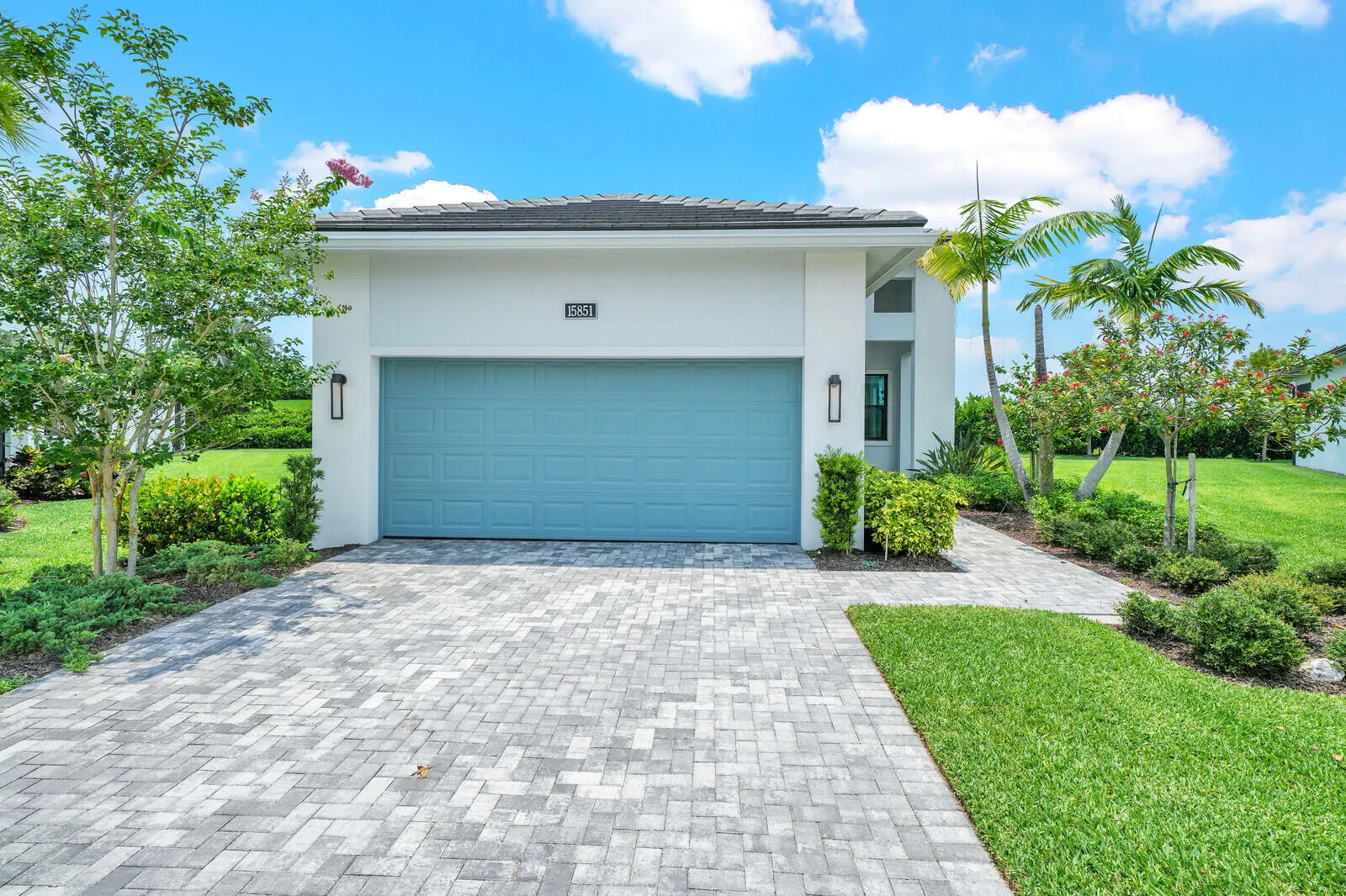 Modern stucco exterior with a tiled roof, featuring a paver driveway, a blue garage door, and lush landscaping
