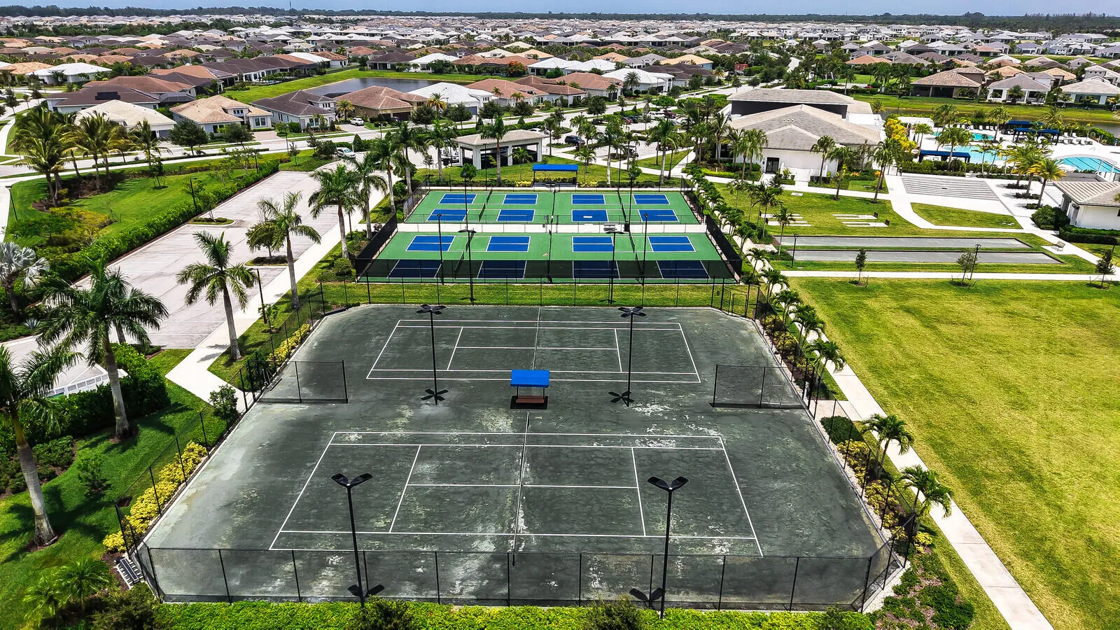 Community amenities featuring multiple tennis courts, pickleball courts, and a manicured lawn area, surrounded by paved pathways and palm trees