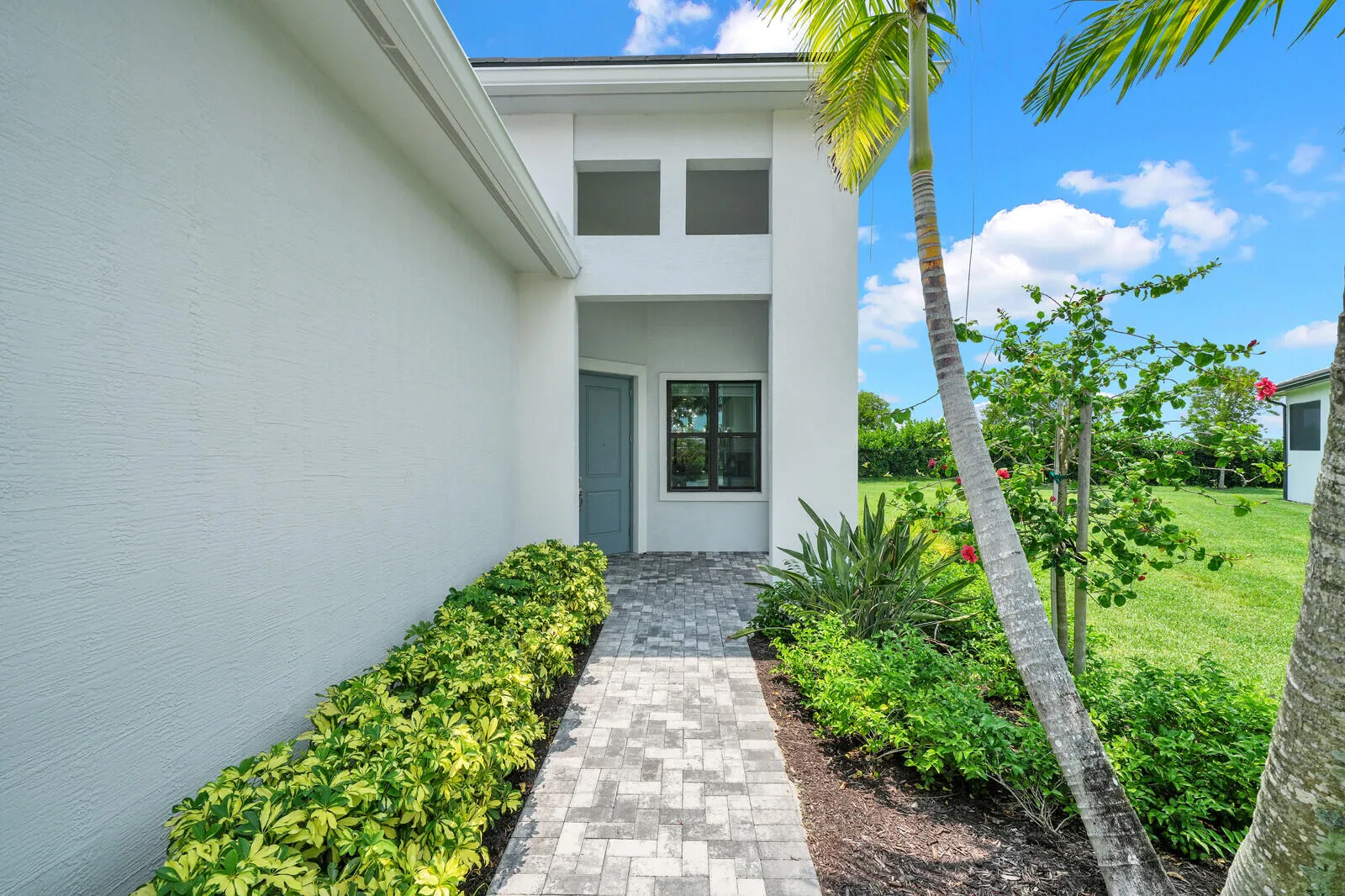 White stucco exterior featuring a recessed entry, paver walkway, mature palm trees, verdant landscaping, and a well-maintained lawn