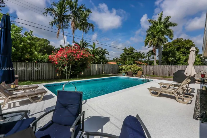 Backyard featuring an in-ground swimming pool, concrete patio, perimeter wood fencing, mature palm trees, and flowering vegetation