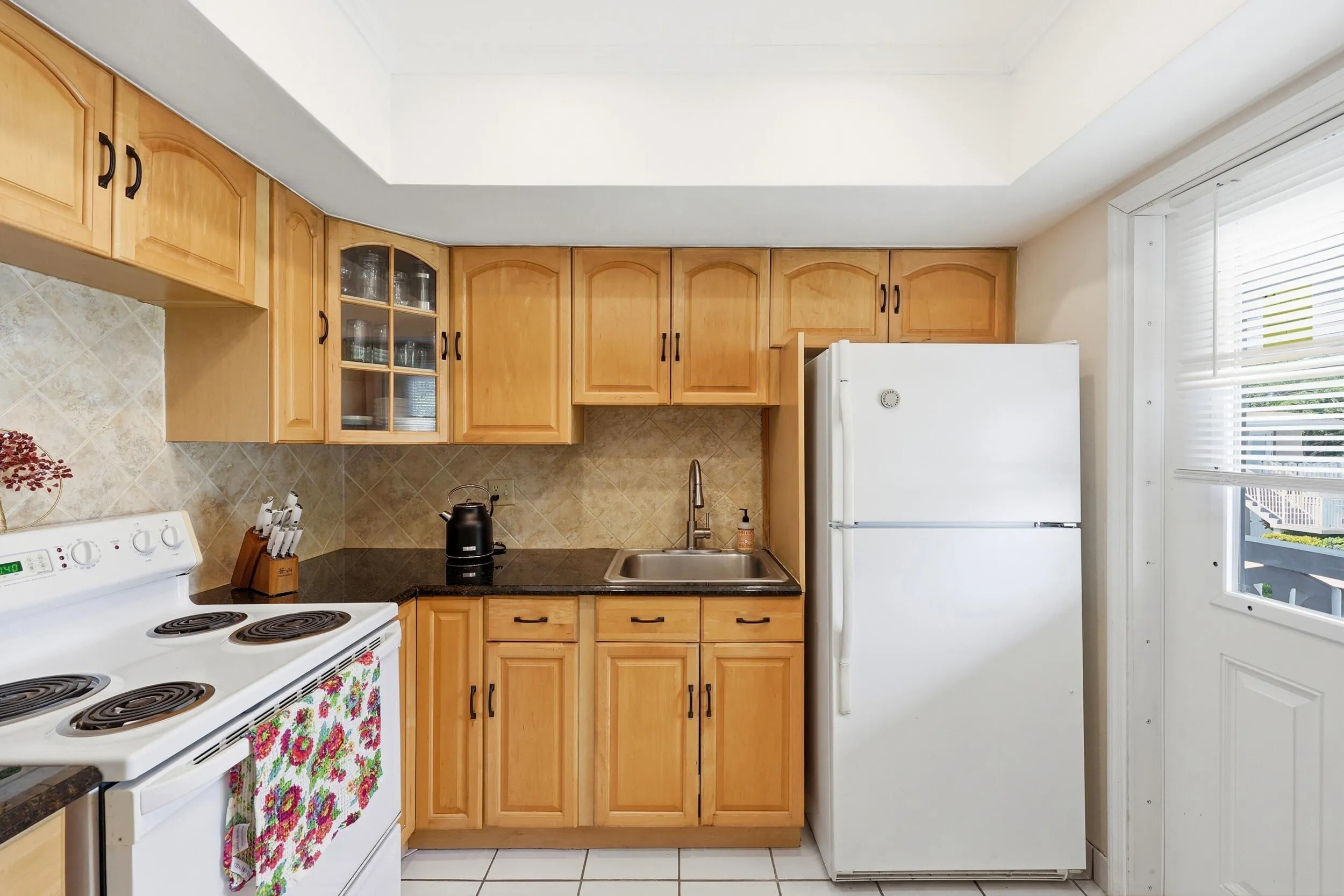 The kitchen offers an efficient and functional layout with abundant cabinet storage, granite countertops, and a neutral tile backsplash that complements the warm wood cabinetry. A tray ceiling enhances the sense of space while ample counter space makes meal preparation easy and convenient. Thoughtful details, including glass-front display cabinetry and additional storage options, provide both practicality and charm. The kitchen also features direct access to the private balcony, allowing natural light to filter in while creating a seamless connection between indoor and outdoor living. A new garbage disposal and new electrical panel were both installed in 2025, adding peace of mind for the next owner.