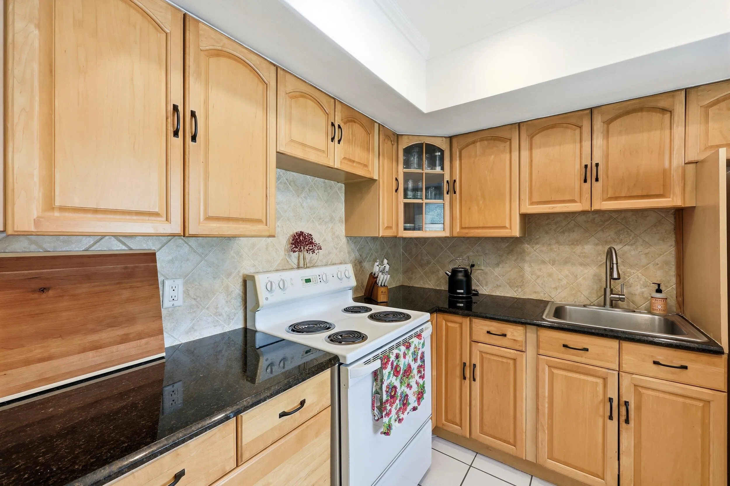 The kitchen offers an efficient and functional layout with abundant cabinet storage, granite countertops, and a neutral tile backsplash that complements the warm wood cabinetry. A tray ceiling enhances the sense of space while ample counter space makes meal preparation easy and convenient. Thoughtful details, including glass-front display cabinetry and additional storage options, provide both practicality and charm. The kitchen also features direct access to the private balcony, allowing natural light to filter in while creating a seamless connection between indoor and outdoor living. A new garbage disposal and new electrical panel were both installed in 2025, adding peace of mind for the next owner.