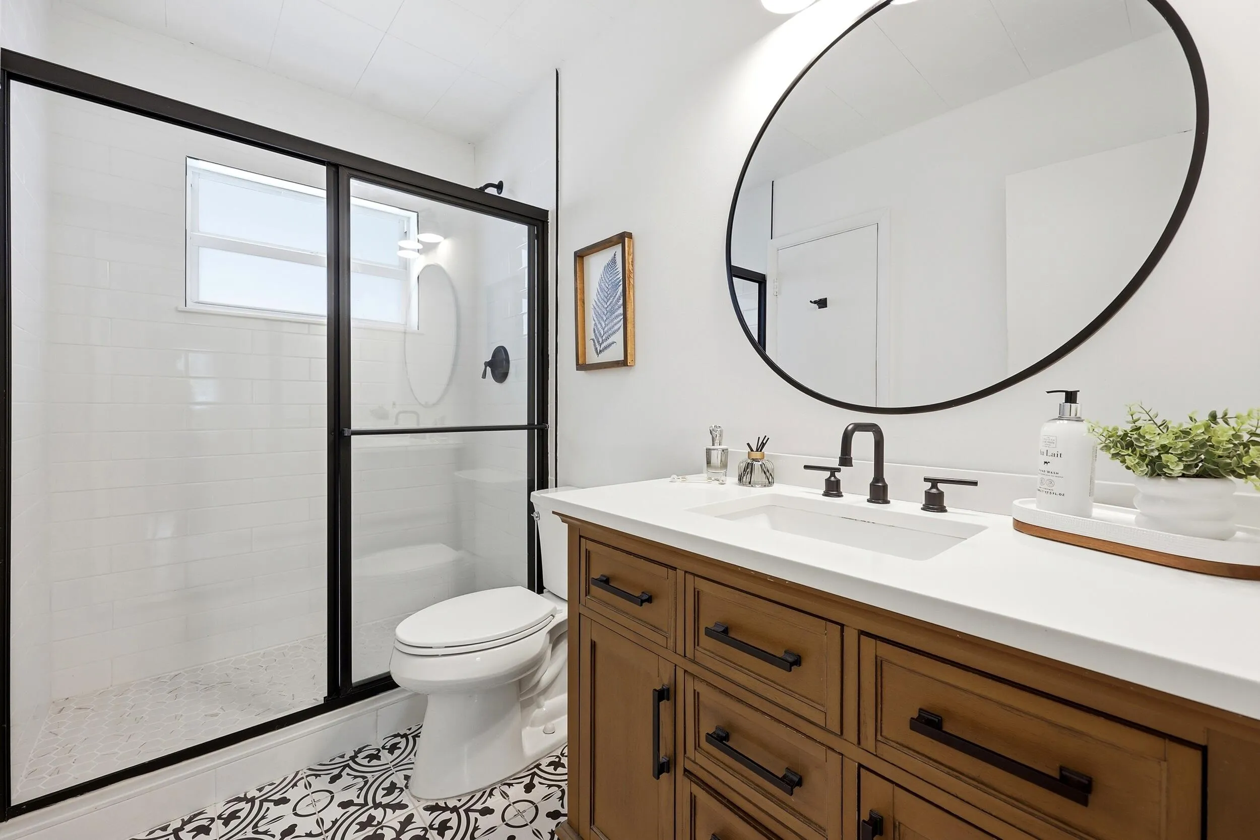 Beautifully renovated, the bathroom blends timeless design with modern functionality. The space features a custom vanity with abundant storage, a quartz countertop, matte black fixtures, designer tile flooring, and a striking round mirror that creates a clean, contemporary feel. The walk-in shower is finished with classic subway tile, a frameless glass enclosure, and a transom window that fills the room with natural light. Thoughtful finishes and quality craftsmanship make this bathroom feel more like a private spa retreat than a typical condominium bath.