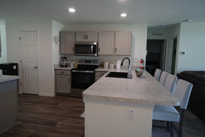 Open-concept kitchen featuring light-toned cabinetry with dark hardware, stainless steel appliances, a center island with an undermount sink and gooseneck faucet, light-toned countertops, and Vinyl plank flooring