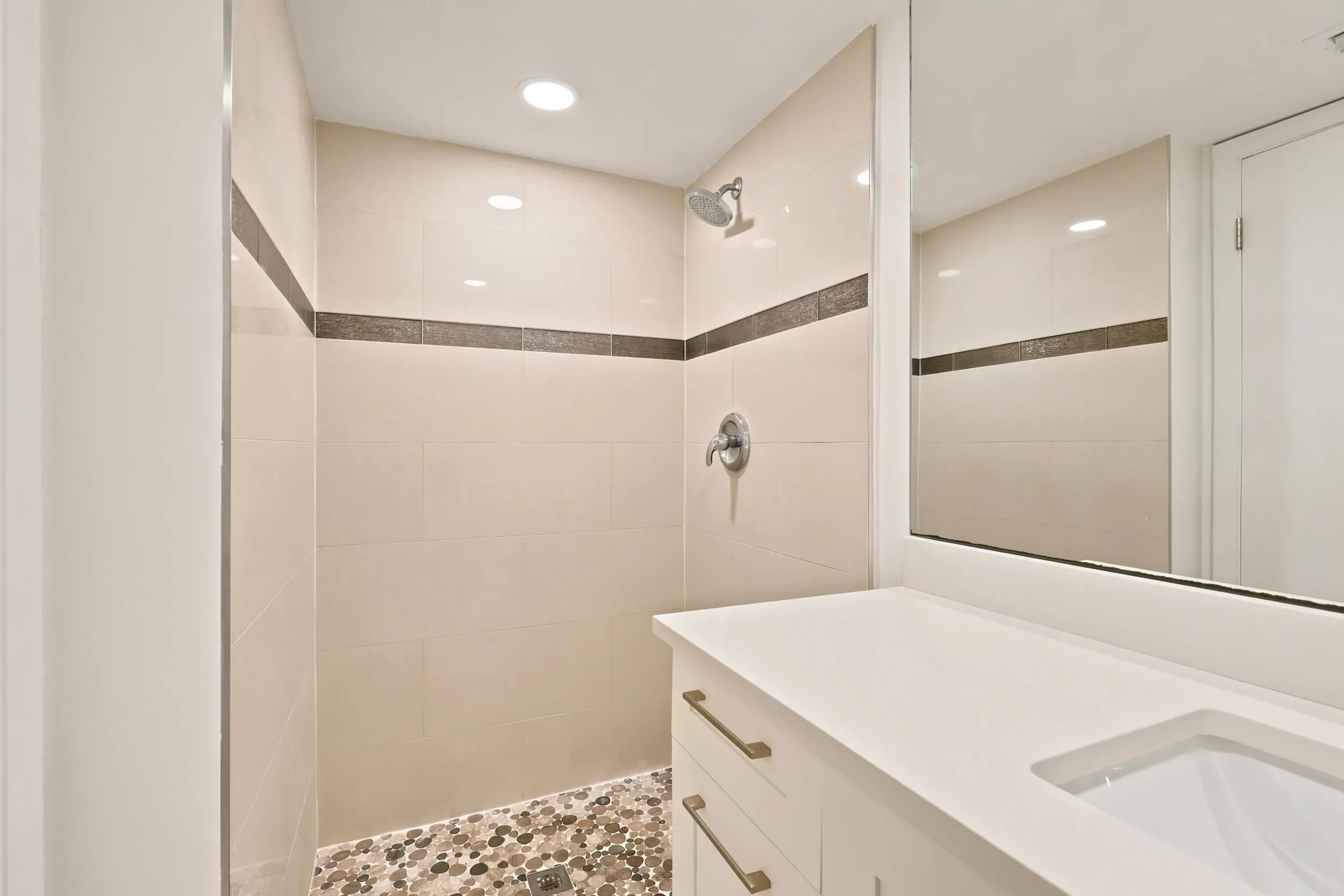 The beautifully renovated primary bathroom showcases a clean, modern design with a sleek white vanity, solid surface countertop, contemporary fixtures, and a large mirror that enhances the sense of space. The walk-in shower features floor-to-ceiling tile, decorative accent detailing, and pebble stone flooring, creating a spa-inspired retreat within the comfort of home.