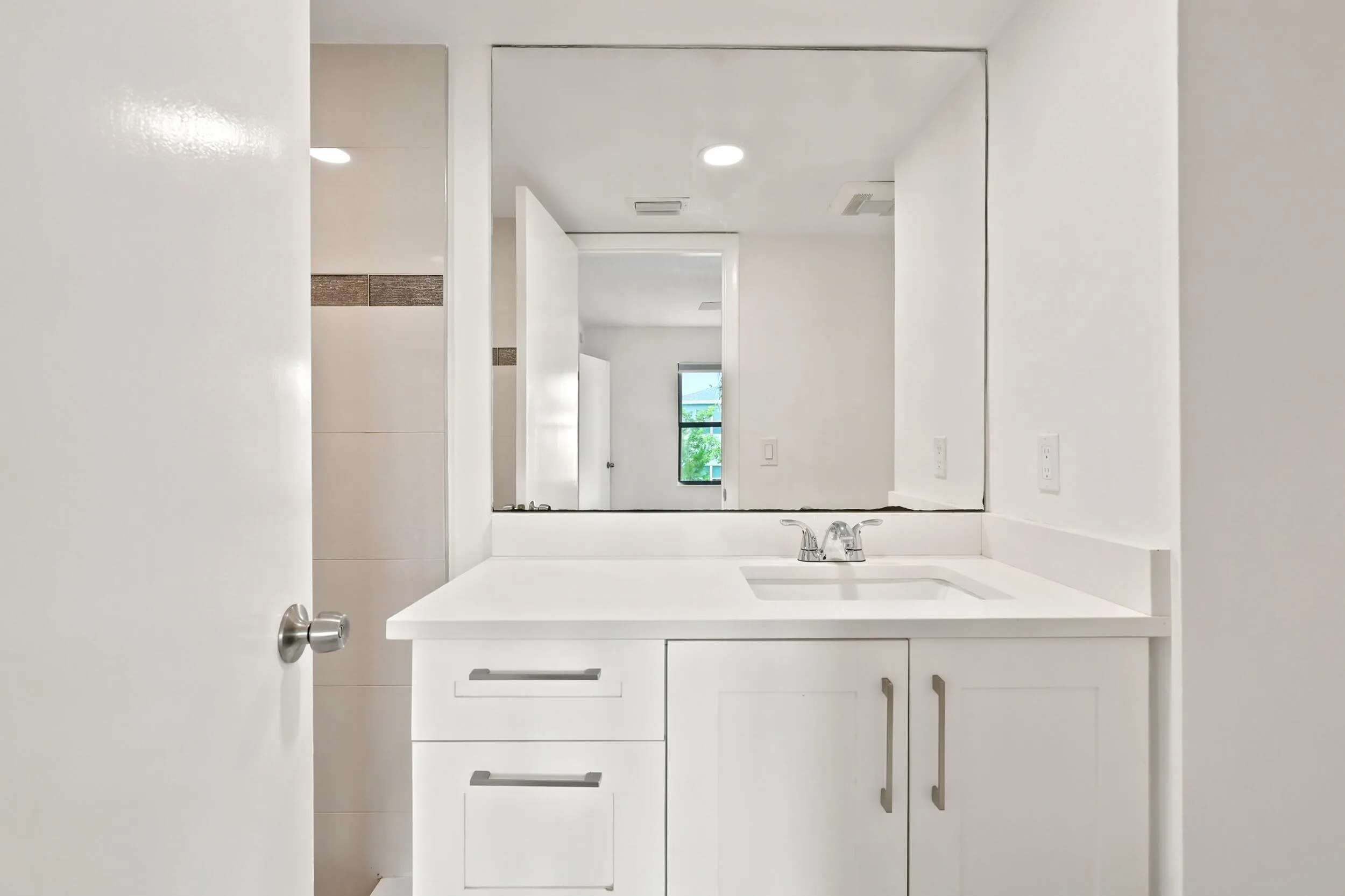 The beautifully renovated primary bathroom showcases a clean, modern design with a sleek white vanity, solid surface countertop, contemporary fixtures, and a large mirror that enhances the sense of space. The walk-in shower features floor-to-ceiling tile, decorative accent detailing, and pebble stone flooring, creating a spa-inspired retreat within the comfort of home.