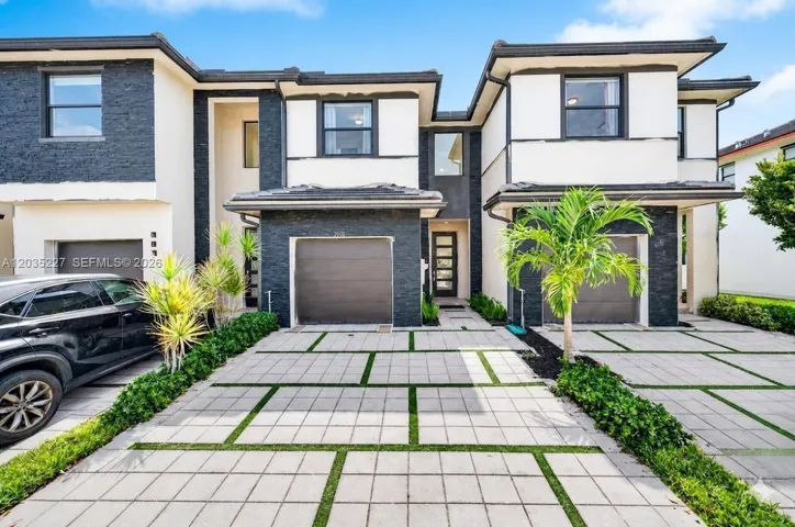 Modern townhome exterior — sleek charcoal stone and white stucco facade, one-car garage, glass-panel entry door, highlighted by landscaping with palms framing a geometric paver driveway.