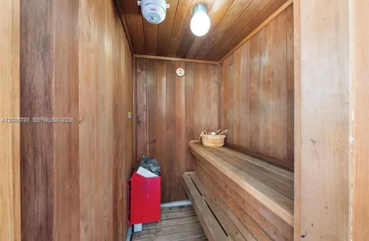 Sauna featuring wood-paneled walls and ceiling, built-in tiered wood bench seating, and a mounted electric heater with stones