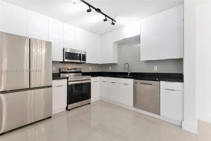 Angled view showcasing the full kitchen layout — floor-to-ceiling white cabinetry, dark granite countertops, full stainless steel appliance suite, and gray subway tile backsplash with polished tile floors.