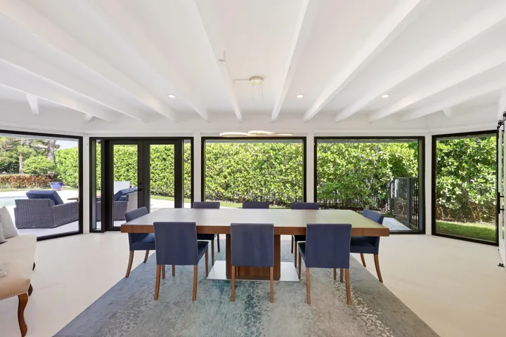 Framed by walls of hurricane impact glass and tranquil views of the pool and lush preserve beyond, the dining room offers an unforgettable setting for everything from intimate dinners to lively gatherings. Exposed wood beams and a striking contemporary chandelier bring warmth, character, and a sense of intimacy to the space, while oversized impact sliders create a seamless connection to the outdoor living areas, allowing the beauty of the surrounding landscape to become part of every meal.