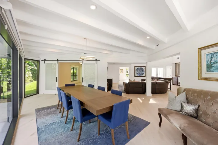 Framed by walls of hurricane impact glass and tranquil views of the pool and lush preserve beyond, the dining room offers an unforgettable setting for everything from intimate dinners to lively gatherings. Exposed wood beams and a striking contemporary chandelier bring warmth, character, and a sense of intimacy to the space, while oversized impact sliders create a seamless connection to the outdoor living areas, allowing the beauty of the surrounding landscape to become part of every meal.