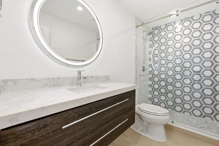 A striking blend of style and sophistication, this beautifully renovated guest bathroom features a custom wood vanity topped with quartz countertops, a designer illuminated mirror, and a frameless glass shower accented by a bold geometric tile feature wall. Contemporary finishes and thoughtful design create a polished space that is both functional and visually impressive for family and guests alike.