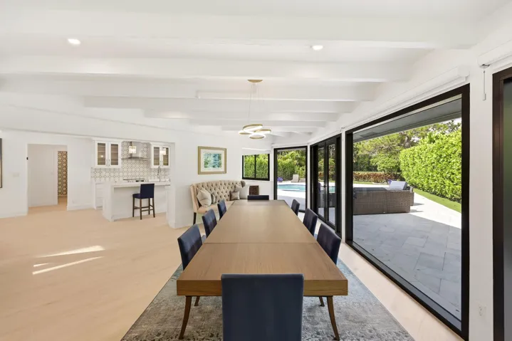Framed by walls of hurricane impact glass and tranquil views of the pool and lush preserve beyond, the dining room offers an unforgettable setting for everything from intimate dinners to lively gatherings. Exposed wood beams and a striking contemporary chandelier bring warmth, character, and a sense of intimacy to the space, while oversized impact sliders create a seamless connection to the outdoor living areas, allowing the beauty of the surrounding landscape to become part of every meal.