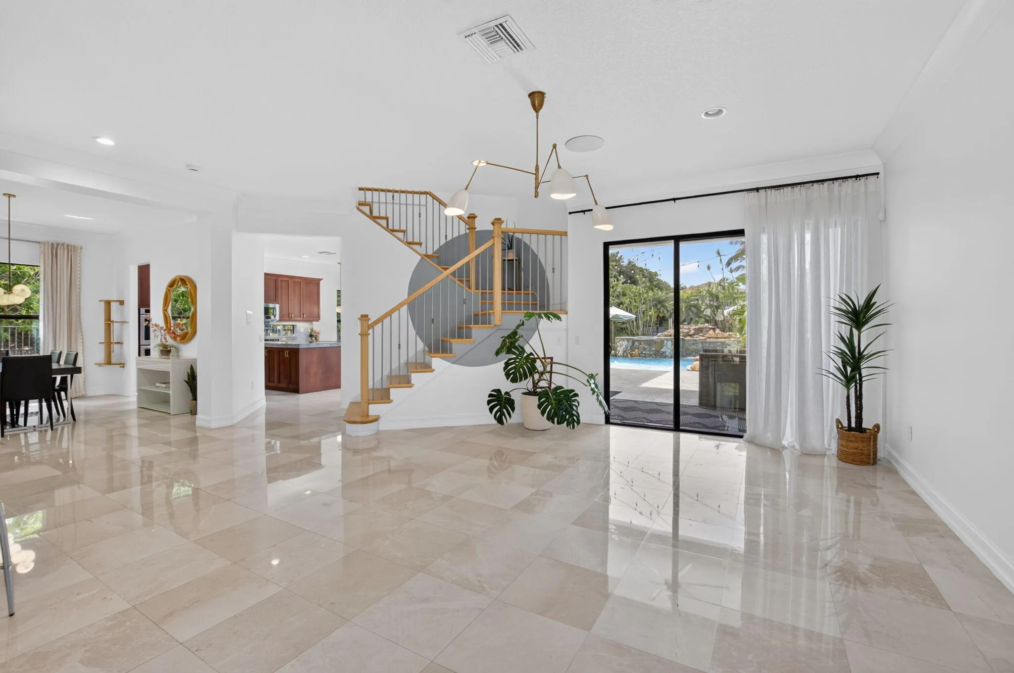 Expansive main living area featuring polished tile flooring, a contemporary chandelier, a staircase with wood newel posts and metal spindles, and large sliding glass doors opening to a pool area