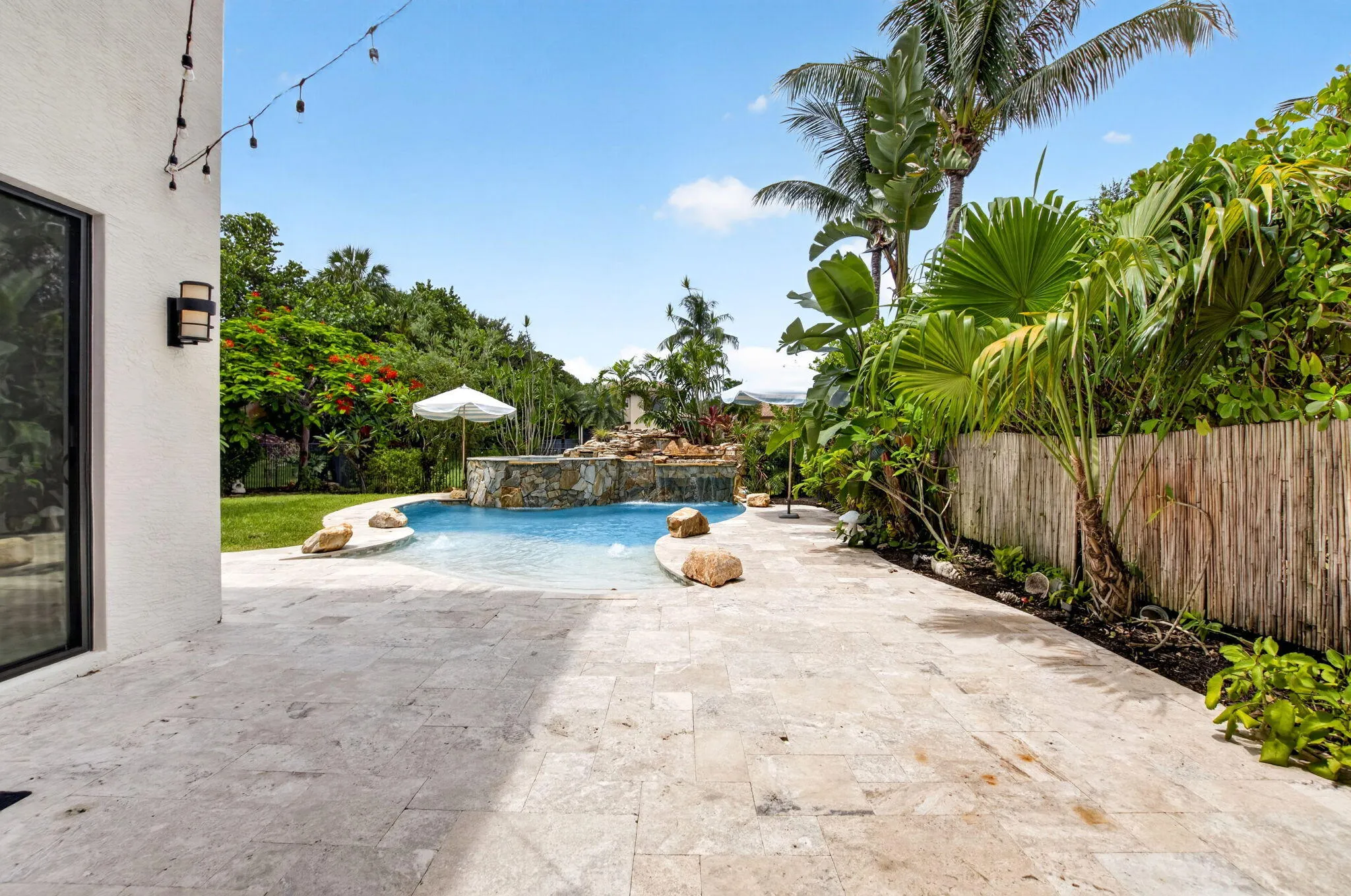 Expansive patio featuring light-toned natural stone pavers, a free-form swimming pool with a rock waterfall feature, tropical landscaping, and a bamboo privacy fence