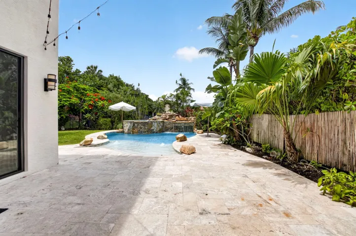 Expansive patio featuring light-toned natural stone pavers, a free-form swimming pool with a rock waterfall feature, tropical landscaping, and a bamboo privacy fence
