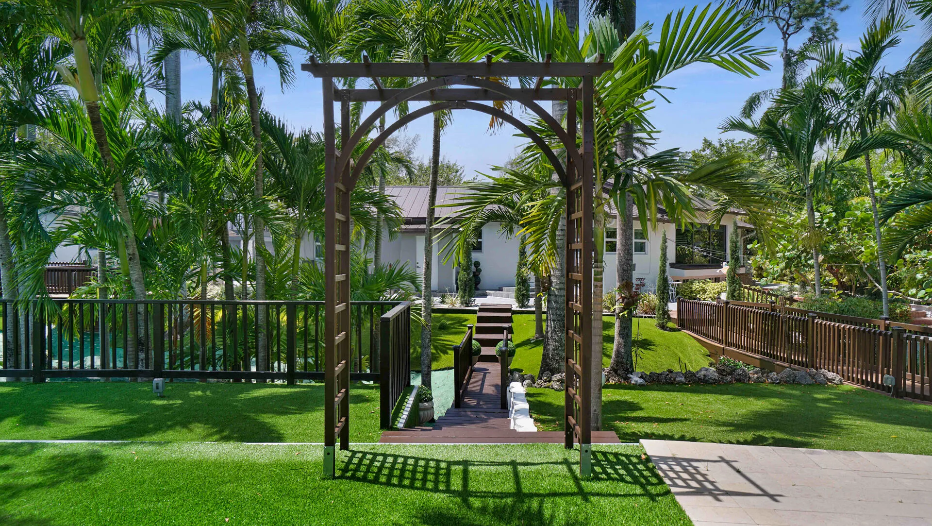 Landscaped exterior featuring a decorative wooden archway, lush green lawn, and mature palm trees