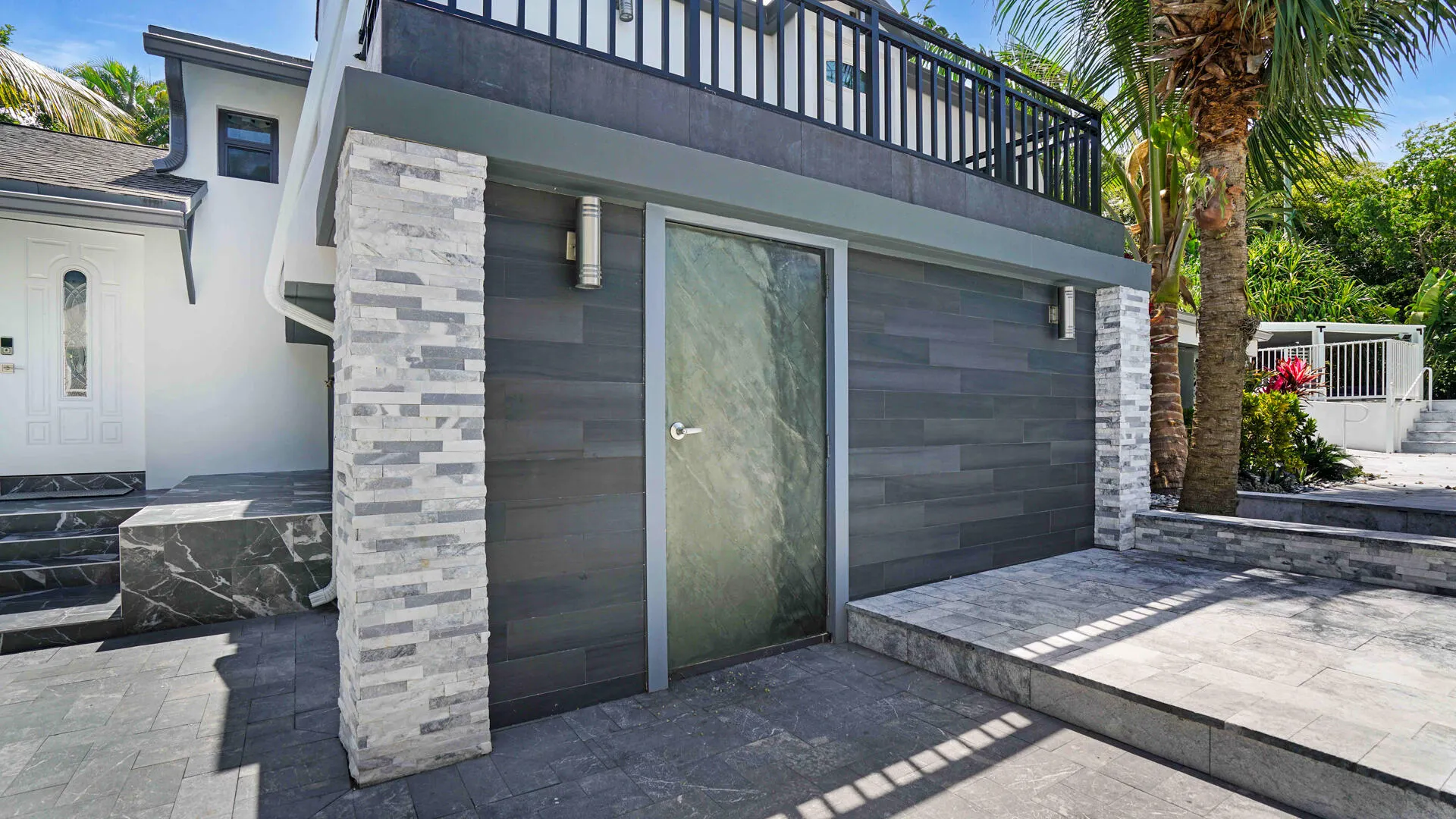 Exterior featuring a balcony with black metal railings, stacked stone columns, a frosted glass entry door, dark gray horizontal siding, and gray paver flooring
