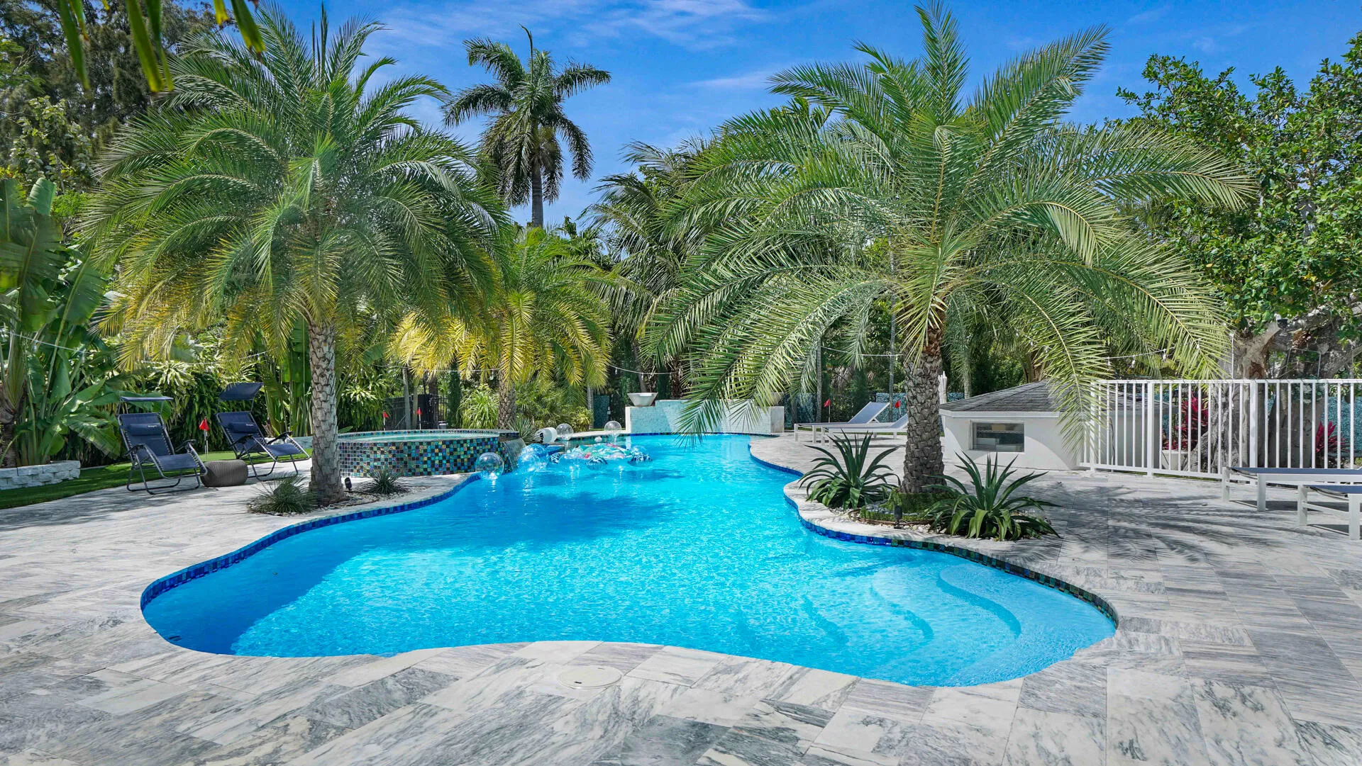Freeform swimming pool with integrated spa and water features, surrounded by extensive patterned paver decking, lush tropical landscaping, and a white picket fence