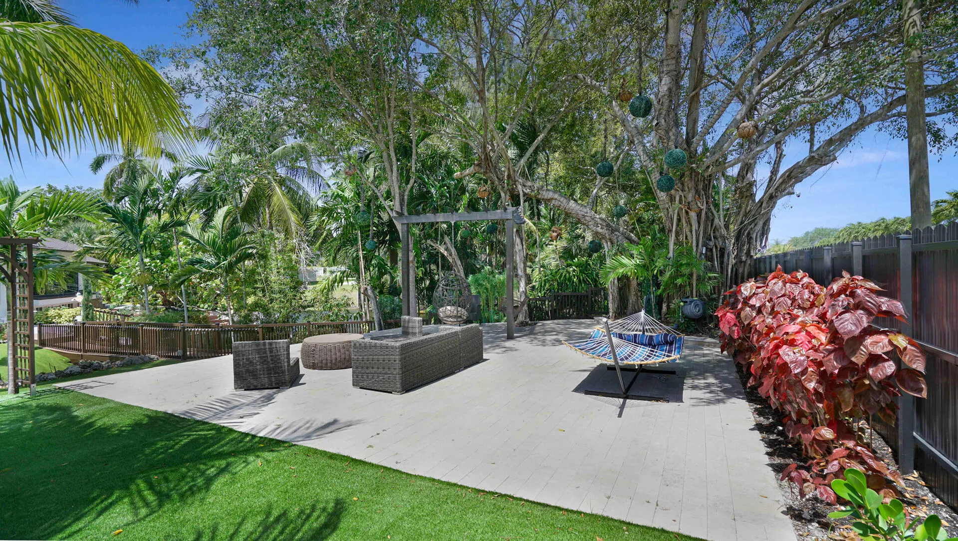 Expansive outdoor living area featuring a large paved patio, artificial turf sections, mature trees providing natural canopy, and lush tropical landscaping