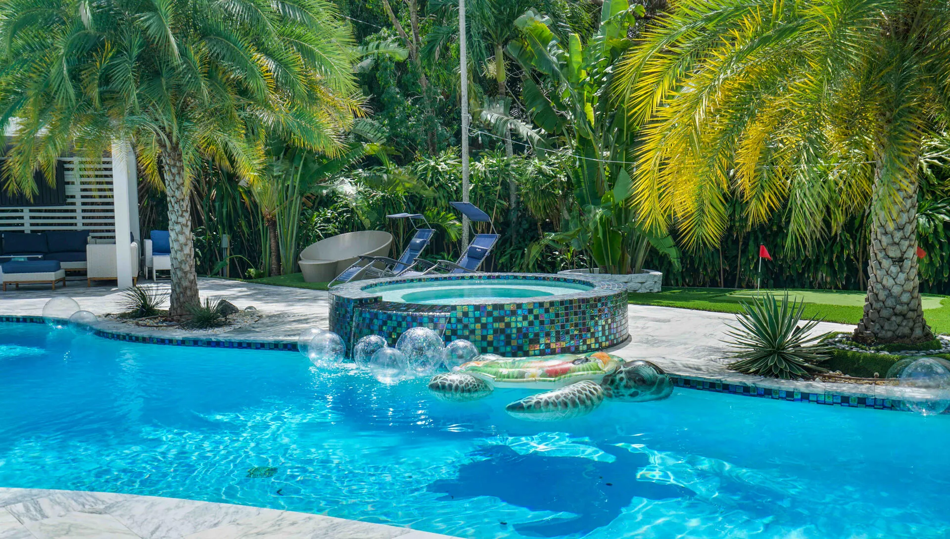 Curved swimming pool with an integrated mosaic-tiled spa, surrounded by tropical landscaping, a covered patio area, and a putting green