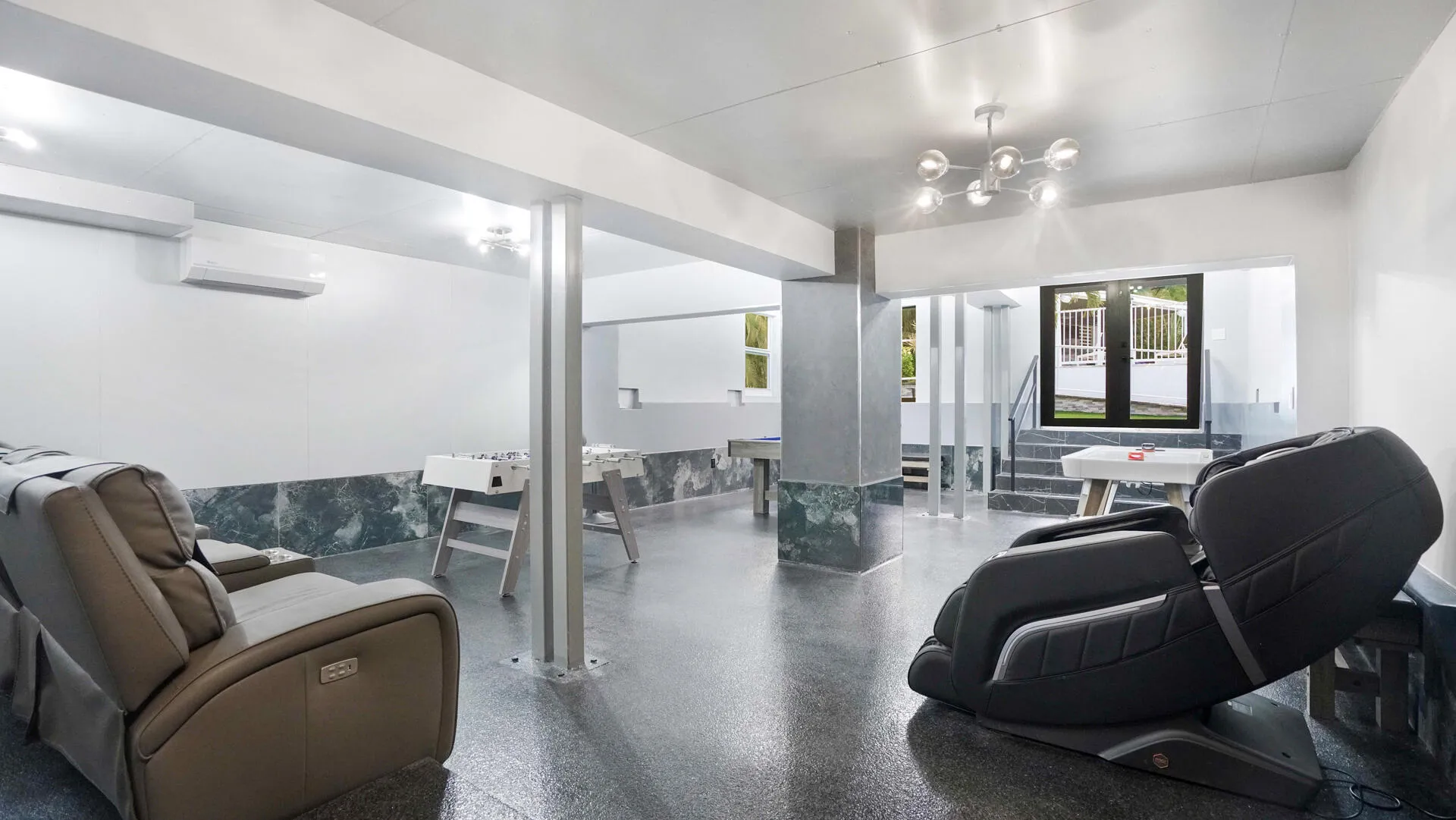 Spacious recreational area featuring polished concrete flooring, a modern multi-bulb light fixture, white walls, and a dark marble-look wainscoting