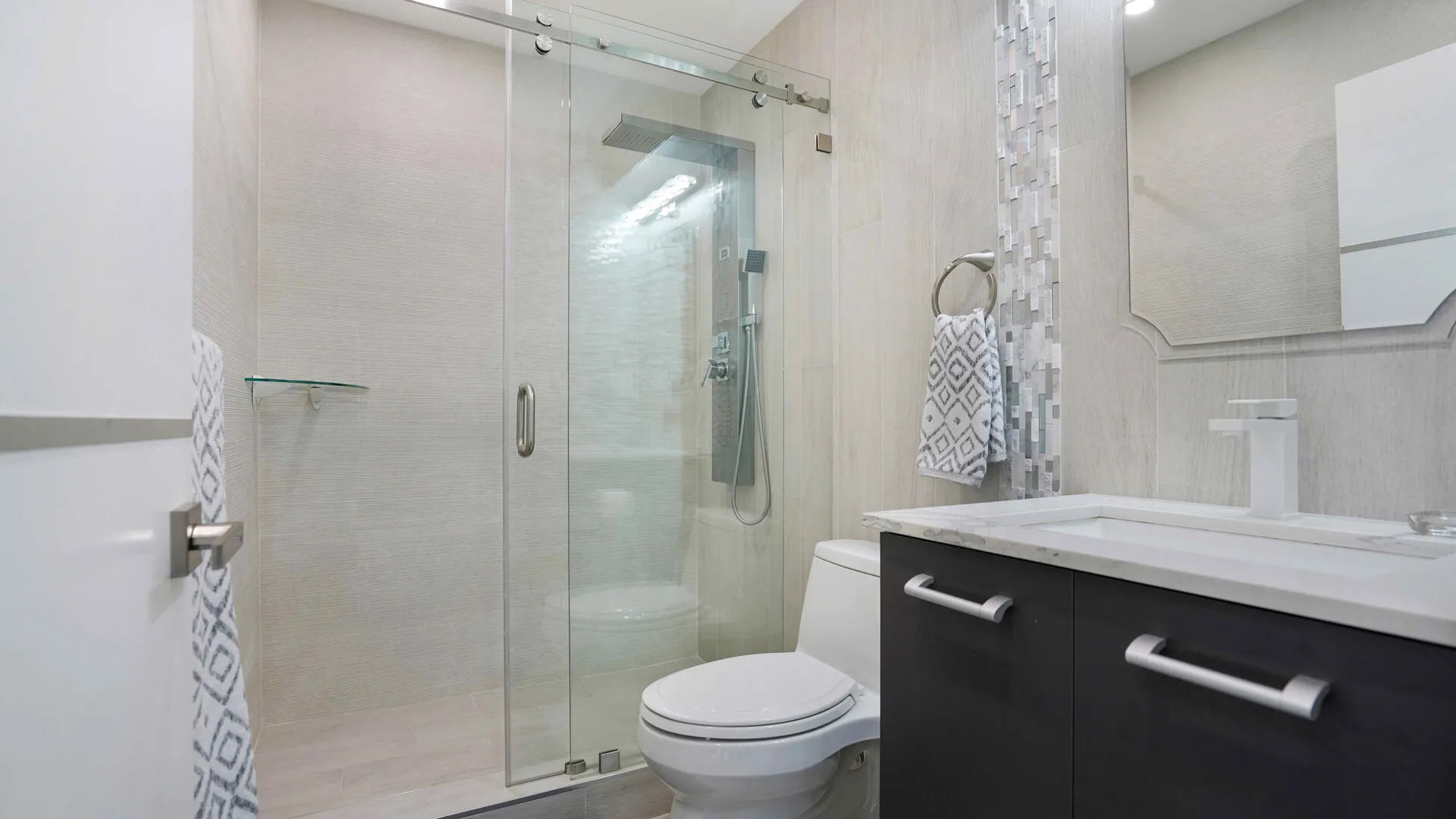 Contemporary bathroom featuring a glass-enclosed shower with a rainfall showerhead, a dark wood-finish vanity with a white countertop and integrated sink, a mosaic tile accent wall, and light-toned wall tiling