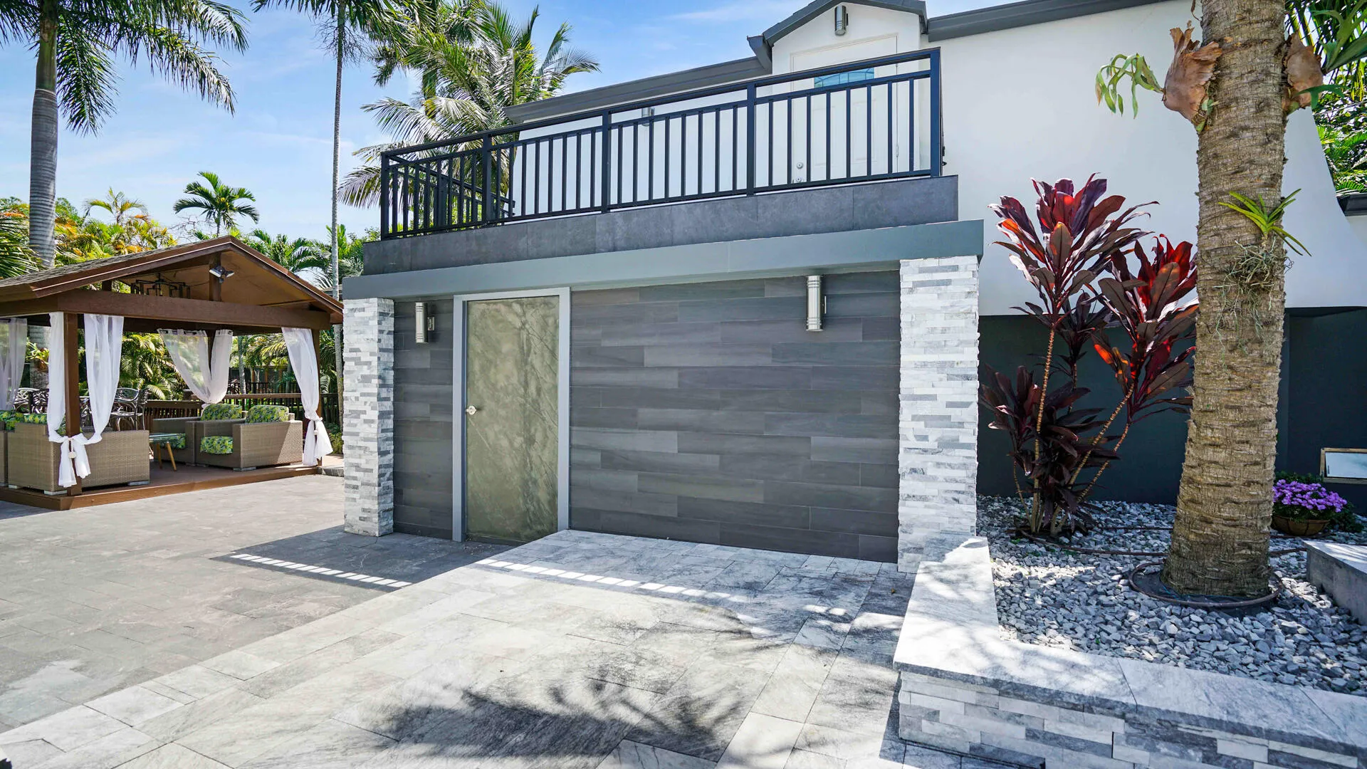 Modern exterior featuring a second-story balcony with dark railings, a ground-level patio with light-toned pavers, and a built-in planter with decorative gravel